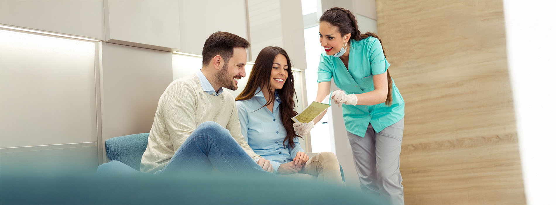 A group of people, including two standing individuals wearing blue scrubs, are gathered around a table with paperwork, smiling and engaged in conversation, with one person holding a clipboard and another sitting on a couch they appear to be in an office setting.
