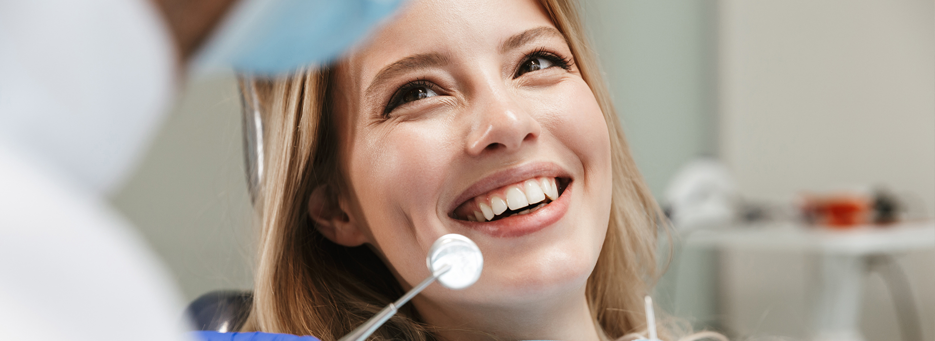 The image shows a woman with a smile, seated in front of a dental professional who appears to be holding a mirror during an examination.